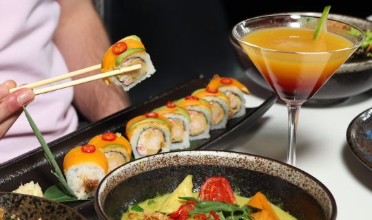Man holding a spicy mango sushi roll with wooden chopsticks over a bowl of vegetable green curry, a cocktail coloured with a red and orange gradient in a martini glass. Crispy chicken salad also in shot.